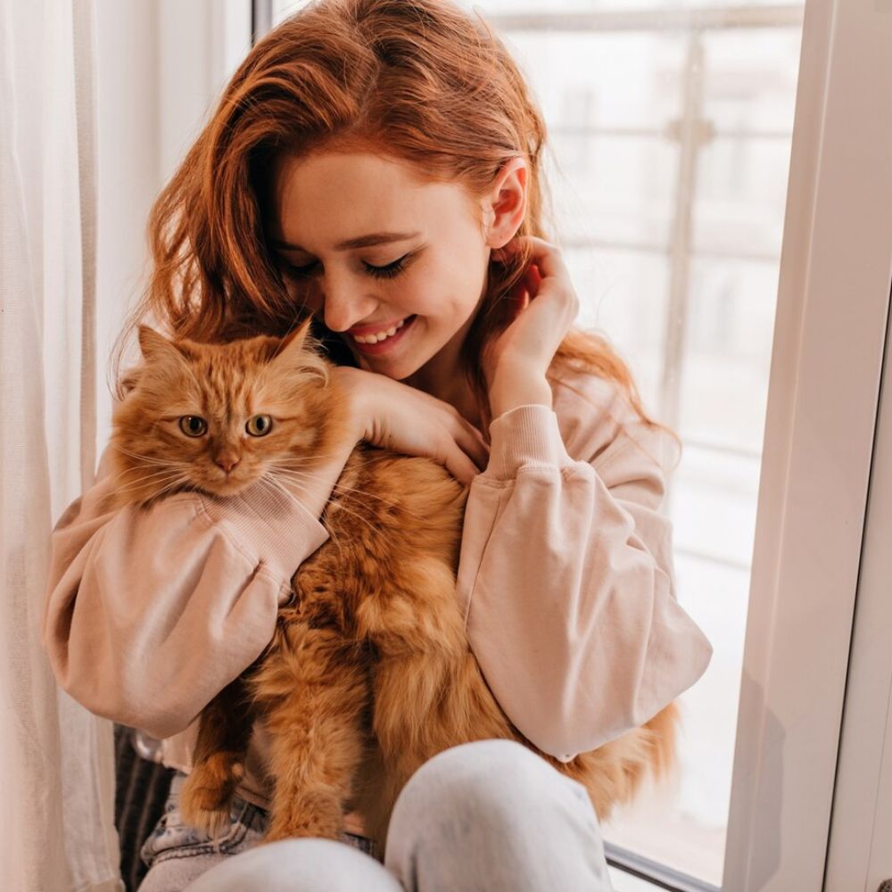 Woman holding a fluffy orange cat in a bright room with light-colored curtains.